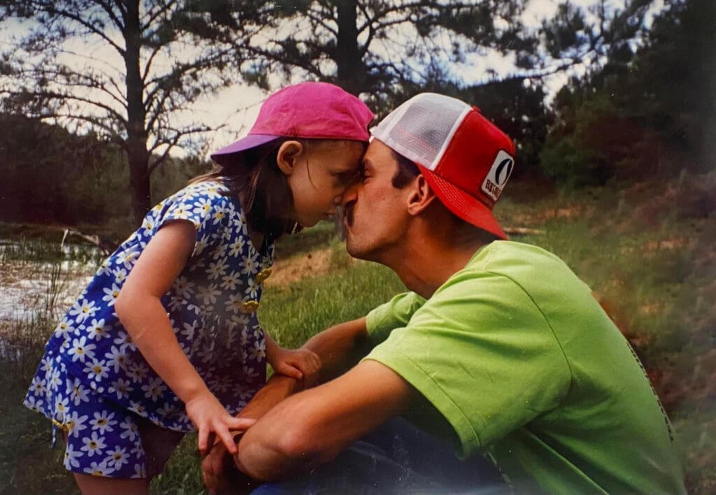 A nostalgic childhood photo of a little girl and her father touching foreheads and sharing a kiss outdoors by a pond, capturing a tender moment of love and connection. This sentimental image represents the importance of taking photos of loved ones and preserving memories that become sacred keepsakes.