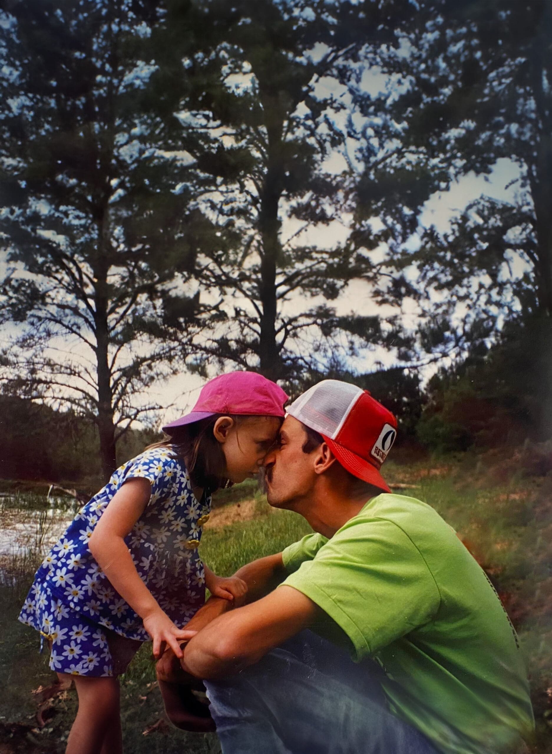A nostalgic childhood photo of a little girl and her father touching foreheads and sharing a kiss outdoors by a pond, capturing a tender moment of love and connection. This sentimental image represents the importance of taking photos of loved ones and preserving memories that become sacred keepsakes.