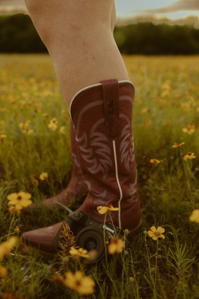 Warm, nostalgic photo of a photographer wearing brown cowboy boots standing in a wildflower field with a camera resting in the grass, symbolizing rooted experience, creative professionalism, and building a strong photography brand
