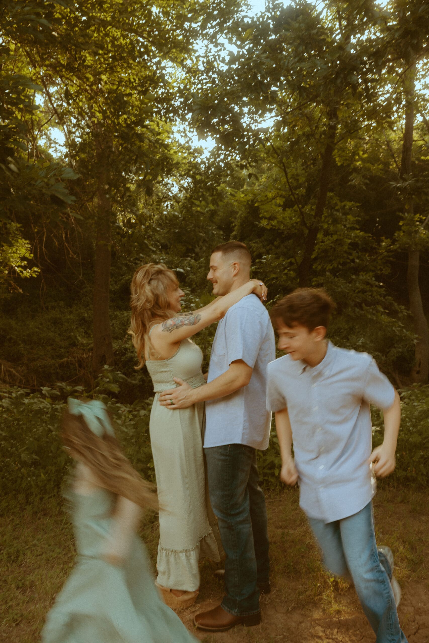 Warm nostalgic family photograph in a sunlit wooded field in Abilene with soft movement blur as children run through the frame while their parents embrace during golden hour. Dreamy storytelling lifestyle photography by Morgan Rose Photography