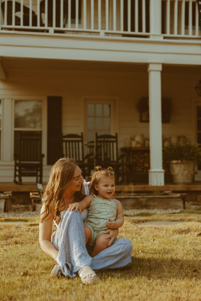 Mother sitting in the grass holding her young daughter in front of a white farmhouse porch during golden hour in Texas. A warm and candid lifestyle motherhood photography moment captured with nostalgic tones and natural emotion by Morgan Rose Photography.