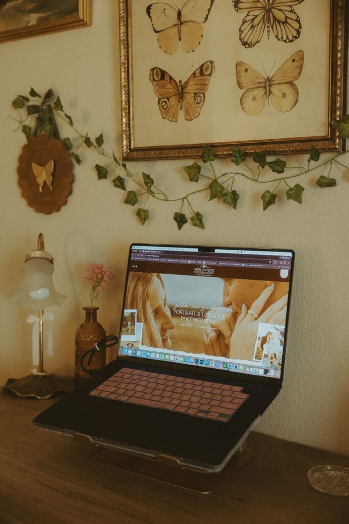 Cozy home office workspace featuring a laptop displaying a Showit photography website, surrounded by vintage butterfly artwork, ivy garland, and warm decor, illustrating website design and branding for photographers comparing Showit vs Pixieset.