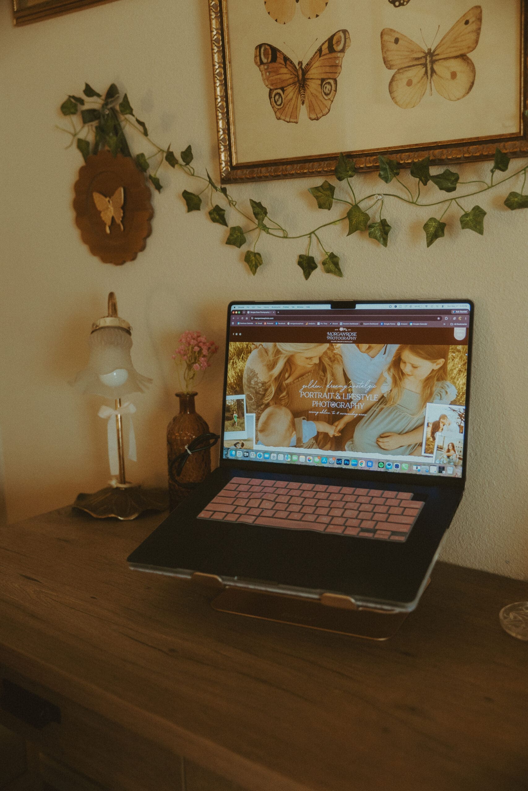 Cozy home office workspace featuring a laptop displaying a Showit photography website, surrounded by vintage butterfly artwork, ivy garland, and warm decor, illustrating website design and branding for photographers comparing Showit vs Pixieset.