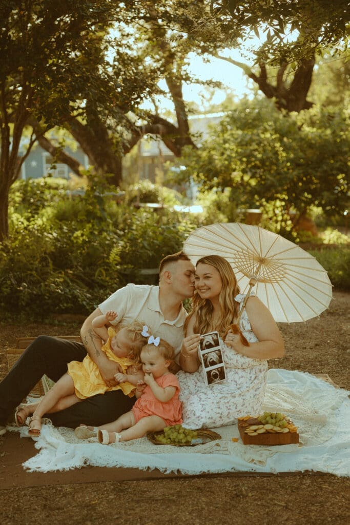 Family sitting together on a picnic blanket during golden hour while celebrating pregnancy with ultrasound photos in a garden setting. A warm and nostalgic lifestyle maternity photography session featuring candid family connection and storytelling moments by Morgan Rose Photography.