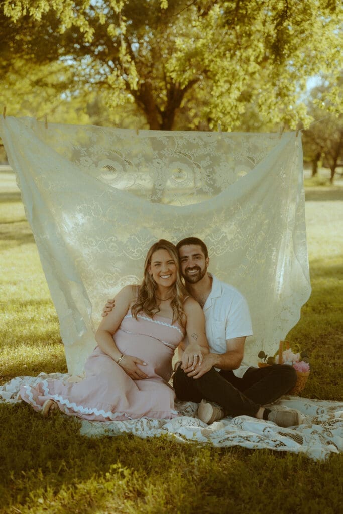Expecting parents sitting together beneath a vintage lace backdrop during golden hour in Abilene surrounded by warm nostalgic light and soft earthy tones. Dreamy maternity storytelling photography by Morgan Rose Photography.