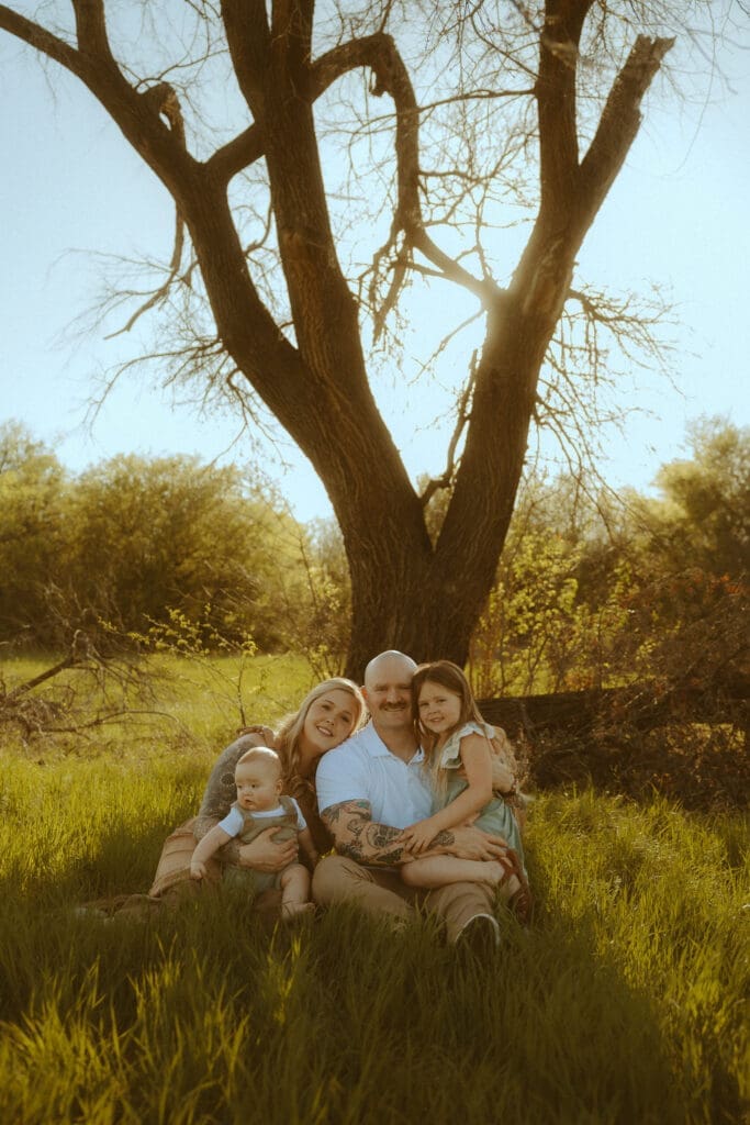 Family of four sitting together beneath a large tree during golden hour in Abilene, Texas, captured in warm natural light during an outdoor family photography session.