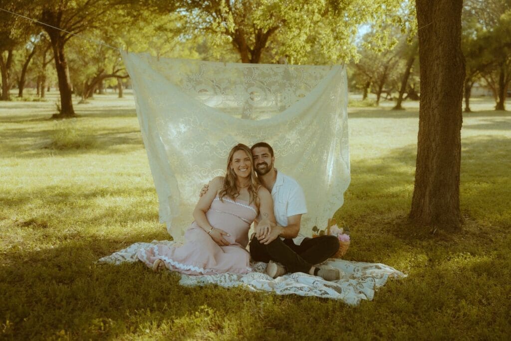 Expecting couple smiling together during a golden hour maternity session in Abilene, Texas, seated on a lace blanket beneath a vintage lace backdrop with warm summer light filtering through the trees.