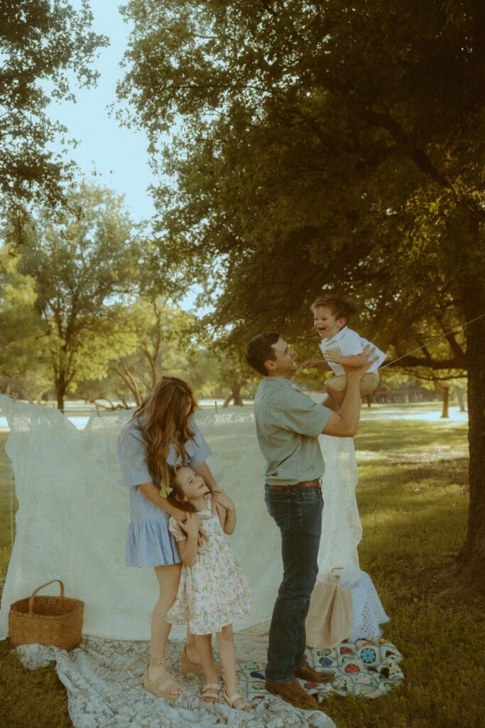 Family enjoying a golden hour summer photo session in Abilene, Texas beneath shaded trees with warm evening light and candid moments between parents and children.