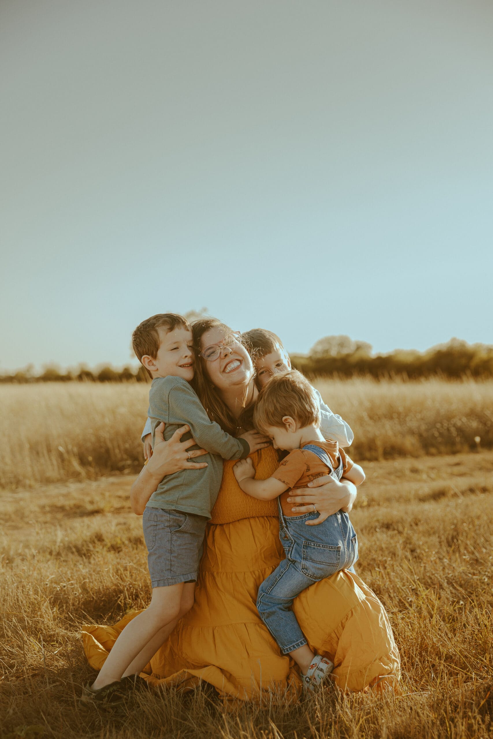 A mother hugs her three young sons during a warm golden hour family photography session in an open field in Abilene, Texas, capturing a genuine moment of love and connection.