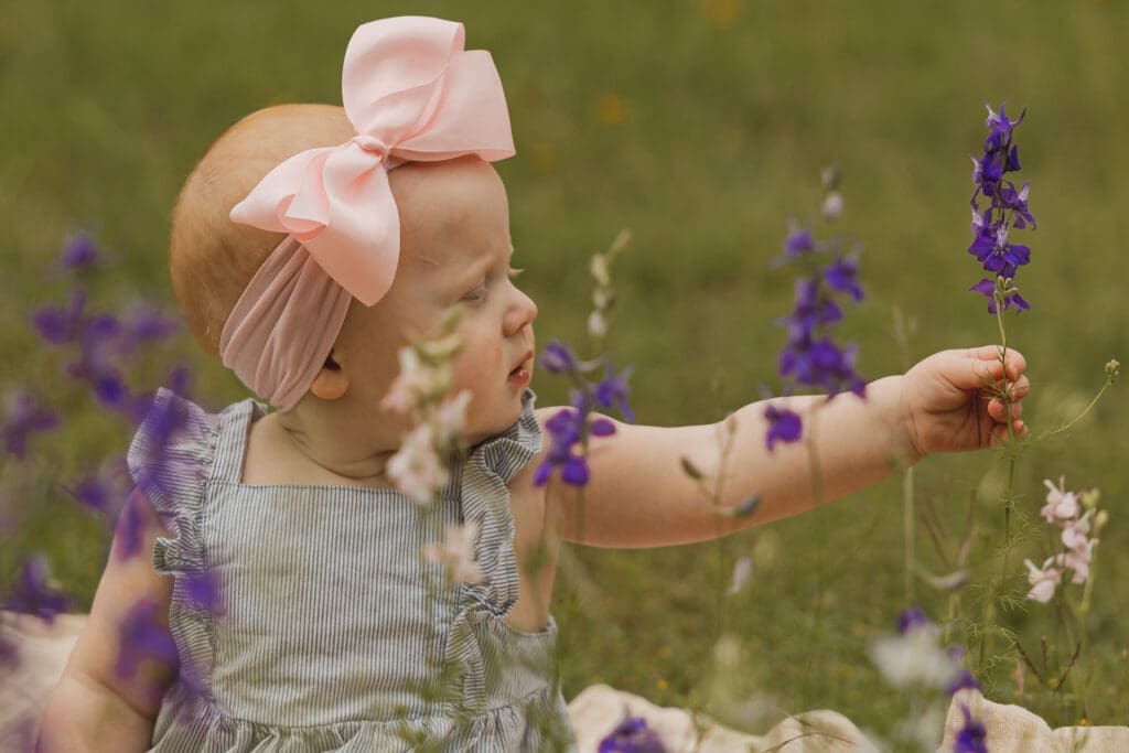 Baby girl reaching out to touch purple wildflowers during an outdoor family photography session in Abilene, Texas, capturing childhood memories through authentic lifestyle photography.