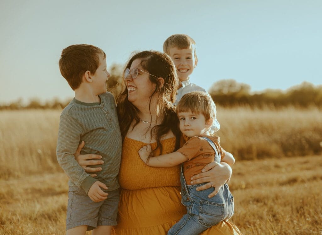 A mother smiles at her young son while holding her three boys during a golden hour family photography session in an open field in Abilene, Texas, capturing a genuine moment of love and connection.