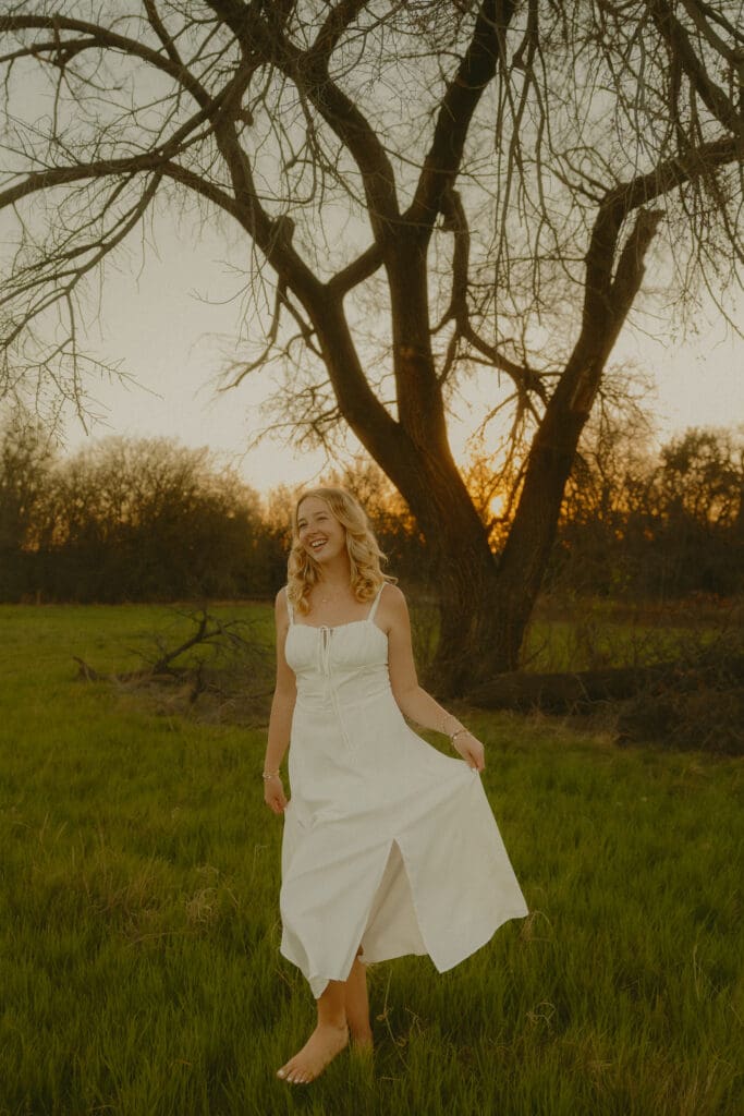 Senior girl smiling and holding her white dress during a golden hour senior photography session in Abilene, Texas, showing a relaxed and natural pose.