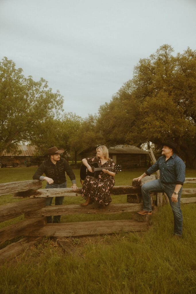 Family of three with an adult son sharing a relaxed moment around a rustic wooden fence, with the mother holding an acoustic guitar during an outdoor family photography session in Abilene, Texas.
